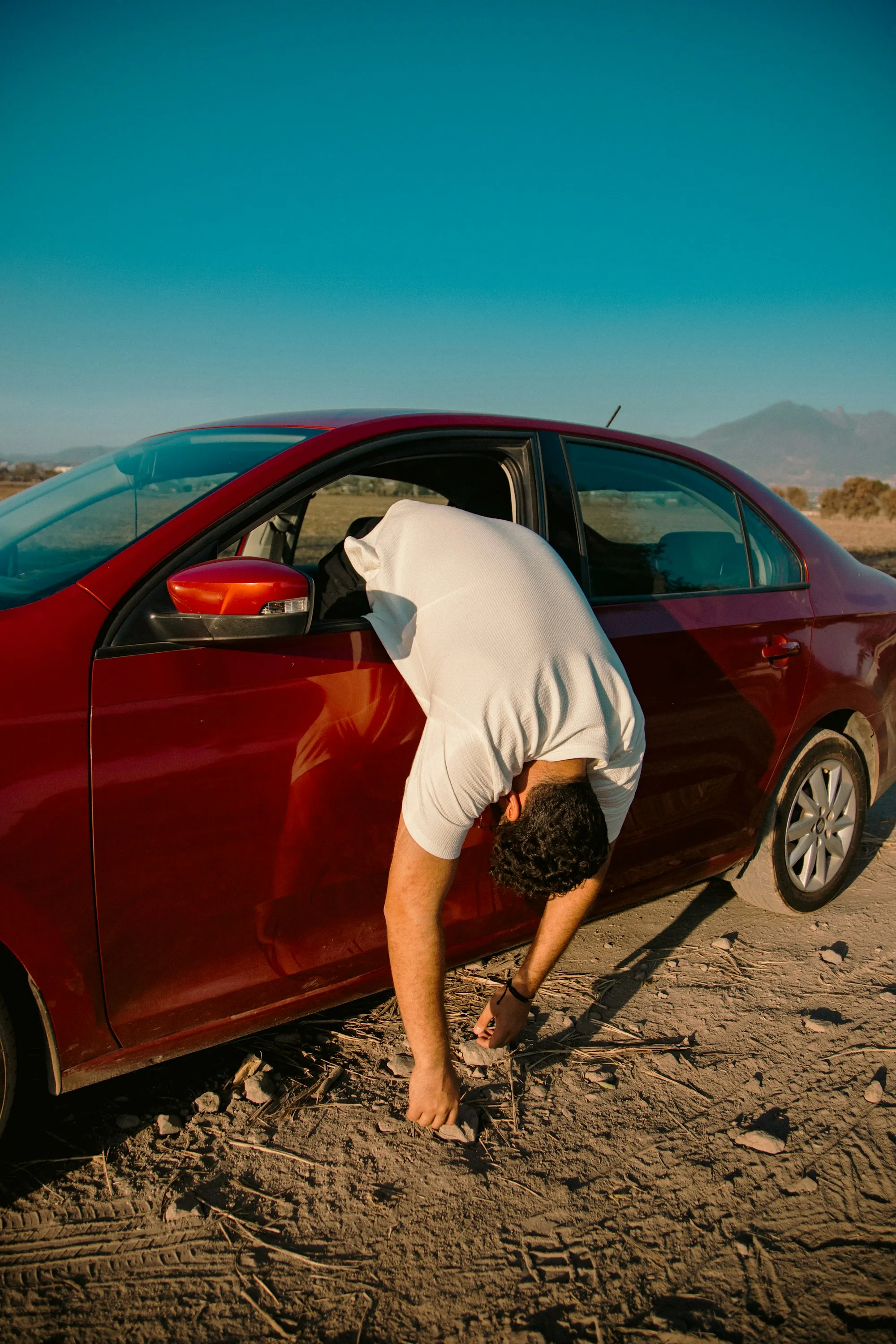 Driver leaning into the open door of a red sedan on a sunny roadside