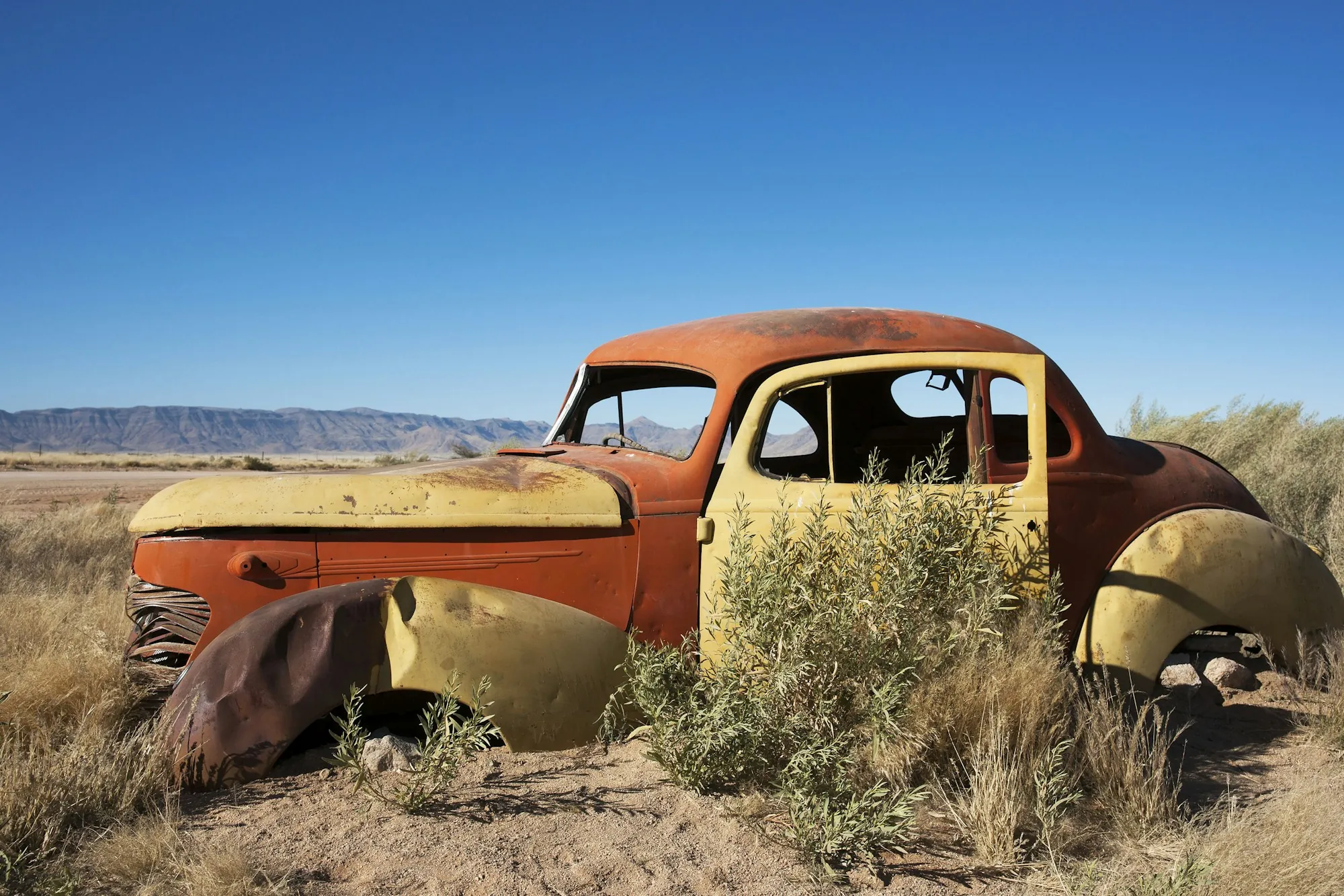 Rusted orange car abandoned in dry brush under a clear sky